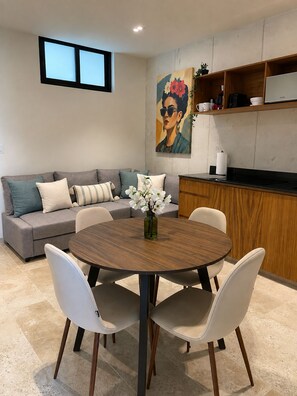Ein moderner Wohnraum mit einem runden Holztisch, umgeben von weißen Stühlen, einem grauen Sofa mit Kissen, einem Bild an der Wand und einer Küchenzeile mit Holzschränken und einer Arbeitsplatte.