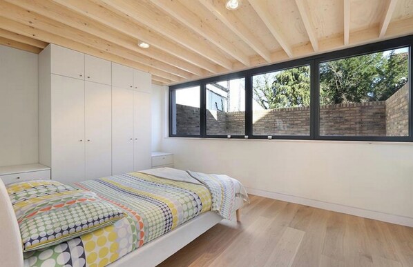 Ein Schlafzimmer mit einem Bett, einem weißen Kleiderschrank und einem Fenster mit Blick auf eine Ziegelwand und Bäume.