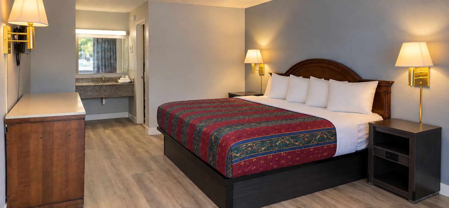 Une chambre d’hôtel comprenant un lit, des tables de chevet, une commode, un meuble en bois et une salle de bain avec un lavabo et un miroir.
