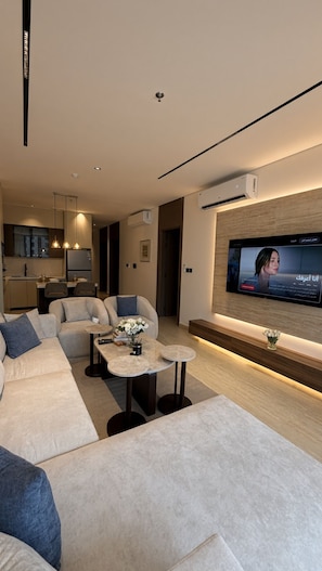 Ein modernes Wohnzimmer mit großem Flachbildfernseher an der Wand, einer Sofagarnitur, einem Couchtisch und einer Küche im Hintergrund.