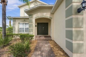 A building with a brick pathway leading to a dark-colored entrance door, surrounded by greenery and a palm tree. - 5BR Splash House in Davenport | Pool & Game Room (Davenport)