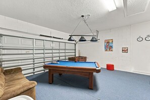 A pool table in a room with a garage door, a red ottoman, and a clock on the wall. - 5BR Splash House in Davenport | Pool & Game Room (Davenport)