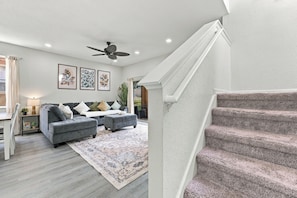 A modern living room with a grey sofa, ottoman, and a patterned rug. A staircase with carpeted steps leads to an upper level. - Regal Palms Haven | 4BR/3.5BA Davenport Resort Townhome with Pool (Davenport)