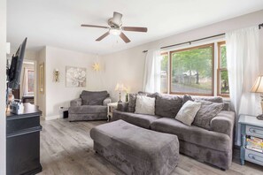 A living room with a grey sofa set, a ceiling fan, a television, and a window with curtains. - Cozy Lakeview Retreat with Patio & Firepit (Rockaway Beach)