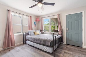 Ein Schlafzimmer mit Bett, Deckenventilator, Fenster mit Vorhängen und einer Tür.