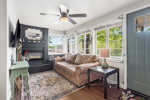 A cozy living room with a fireplace, a sofa, a coffee table, and a view of the outdoors through large windows. - The Ravens Roost Lakeview Retreat-Walk to the Park/Fishing/Boating (Rockaway Beach)