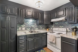 A kitchen with dark cabinets, a white stove, and a patterned backsplash. - The Ravens Roost Lakeview Retreat-Walk to the Park/Fishing/Boating (Rockaway Beach)