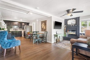 Un salon moderne avec une cheminée, un coin repas avec une table et des chaises, et une cuisine dotée de meubles sombres et d’un dosseret carrelé.