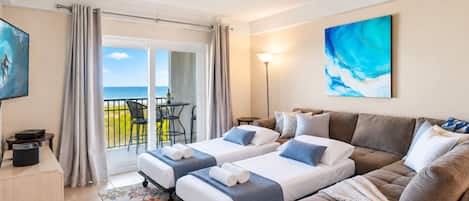 A hotel room with two beds, a sofa, a TV, a desk, and a balcony with a view of the sea.