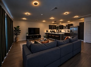 A modern living room with a large sofa, a flat-screen TV, and a kitchen area with dark cabinetry and a central island. - Heights Gem Chef's Kitchen 5 Min to Downtown (Houston)