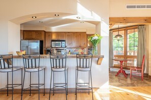 A kitchen with a central island, bar stools, wooden cabinets, a refrigerator, a microwave, and a window with curtains. - Equinox Condo | Ski-in/Ski-out  | Stunning Lake & Mountain View | Pool Access (Mont-Tremblant)