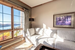 A living room with a large window, a sofa, a lamp, and a framed picture on the wall. - Equinox Condo | Ski-in/Ski-out  | Stunning Lake & Mountain View | Pool Access (Mont-Tremblant)