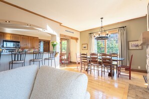 A spacious living area with a sofa, a dining table with chairs, a kitchen with a microwave, and a large window with curtains. - Equinox Condo | Ski-in/Ski-out  | Stunning Lake & Mountain View | Pool Access (Mont-Tremblant)