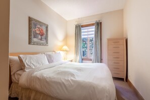 Ein ordentlich bezogenes Bett mit weißen Leinentüchern, eine Nachttischlampe, ein gerahmtes Blumenbild an der Wand und ein Holzschrank mit mehreren Schubladen.