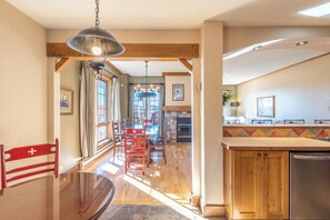 A spacious dining area with a wooden table and chairs, a kitchen with a stone backsplash, and a view of the outdoors. - Equinox Condo | Ski-in/Ski-out  | Stunning Lake & Mountain View | Pool Access (Mont-Tremblant)