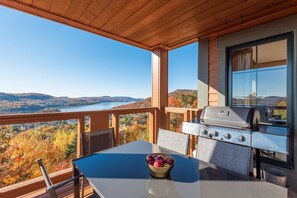 A balcony with a view of a lake and hills, a grill, and a table with a bowl of fruit. - Equinox Condo | Ski-in/Ski-out  | Stunning Lake & Mountain View | Pool Access (Mont-Tremblant)
