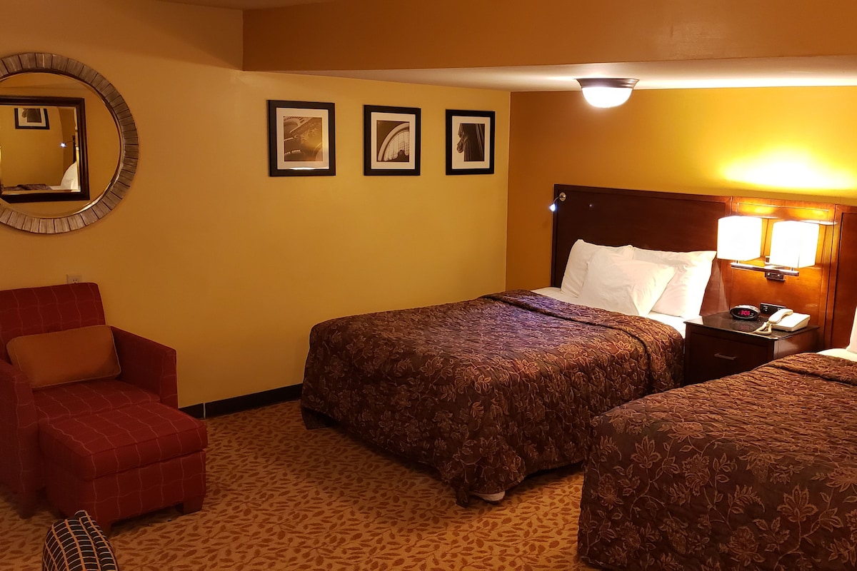 A hotel room with two beds, a red armchair, a round mirror, and three framed pictures on the wall.