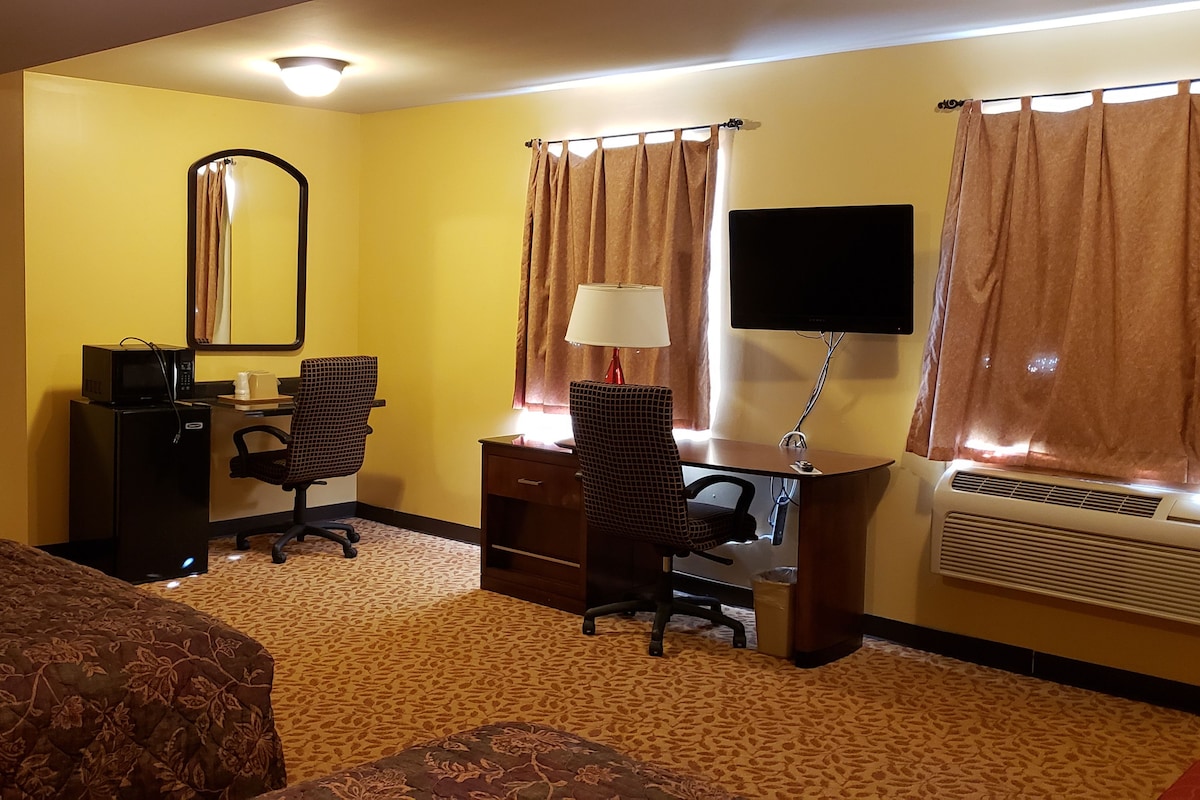 A hotel room with a bed, a desk with a chair, a mirror, a television, and an air conditioning unit.