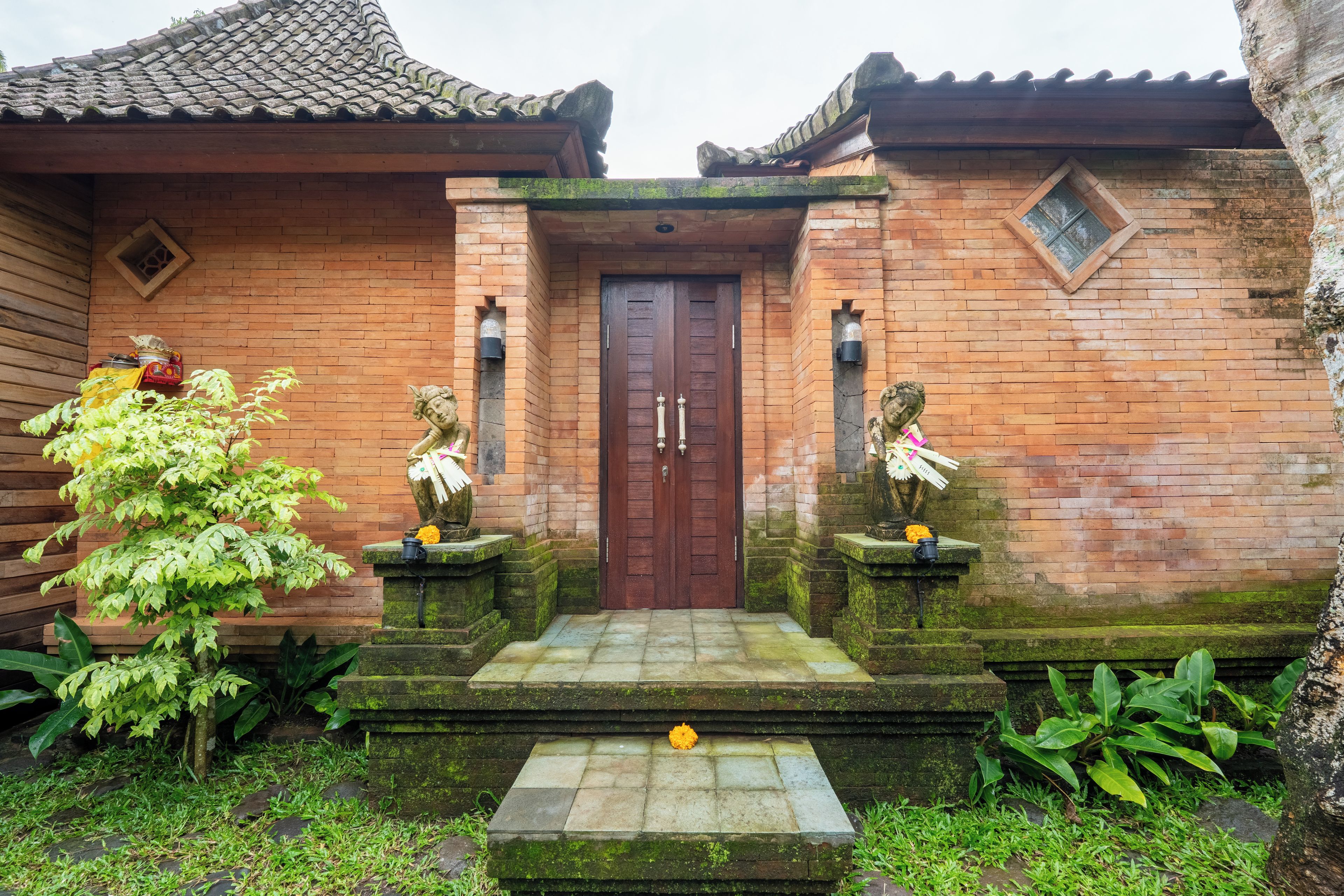 A traditional brick building with a tiled roof, a wooden door, and two stone statues on either side of the entrance.