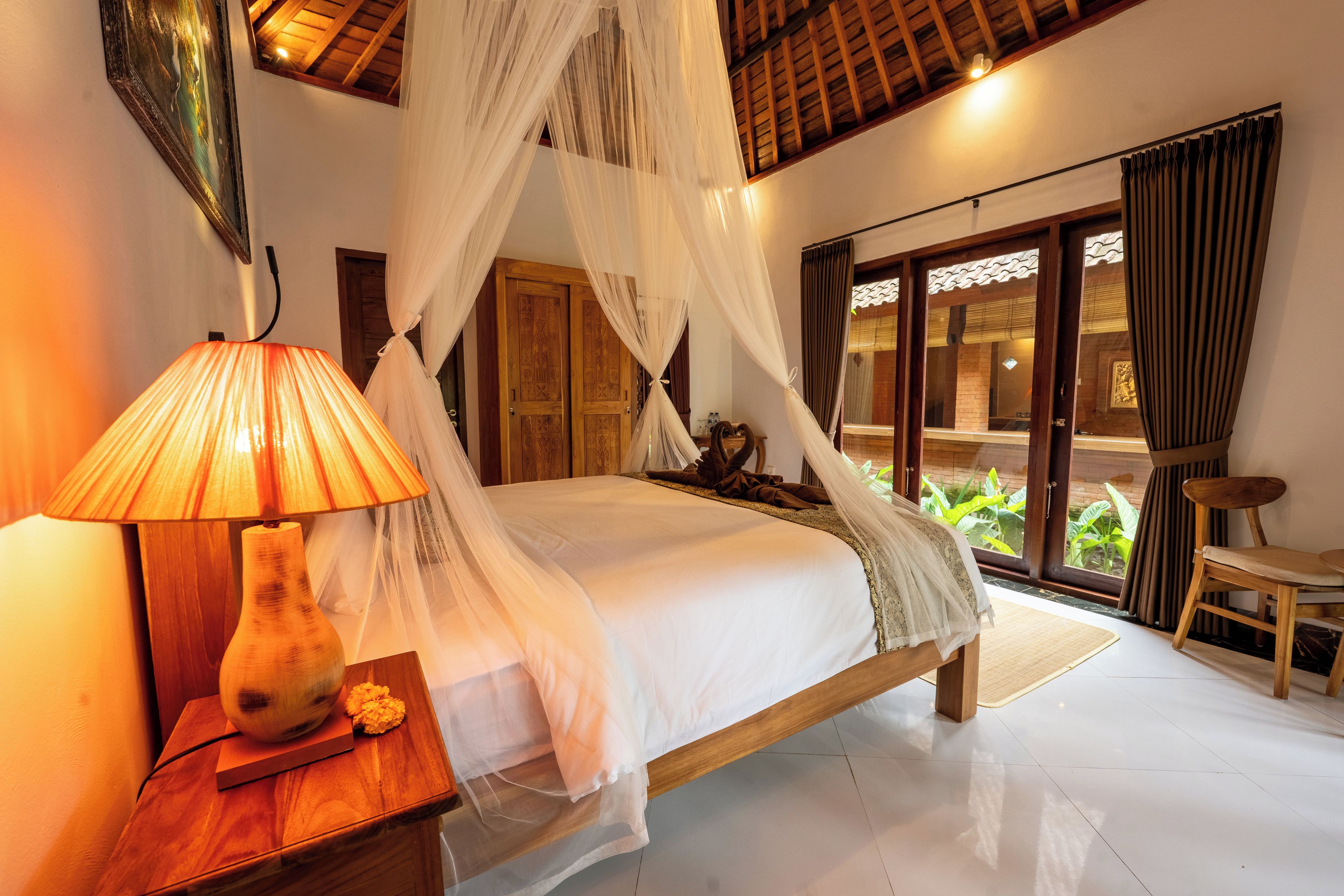 A bedroom with a bed, a bedside lamp, a wooden nightstand, and a view of greenery through a glass door.