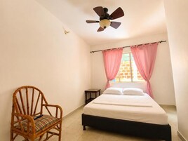 Un dormitorio con una cama, una silla, una mesita de noche, un ventilador de techo y cortinas rosadas.