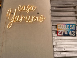 Un letrero de neón con la frase 'casa Yarum' y una señalización callejera colorida que indica 'CALLE 51 27-43'.