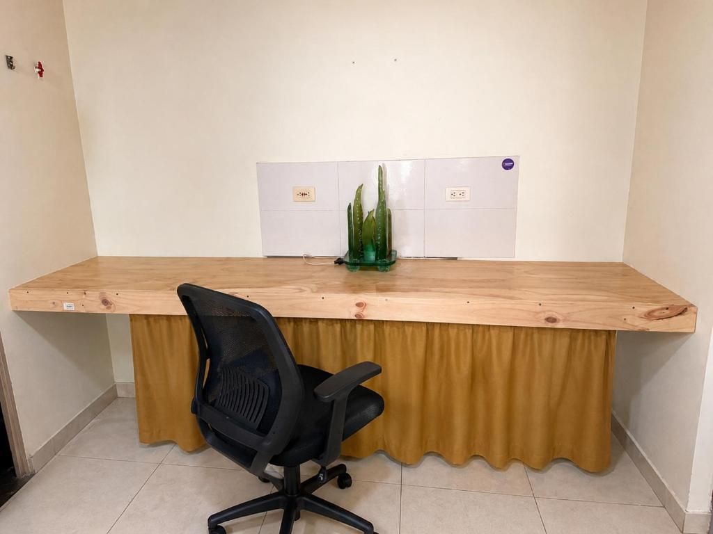 Un escritorio de madera con una silla de oficina negra, una planta verde y una pared de azulejos blancos con un enchufe.