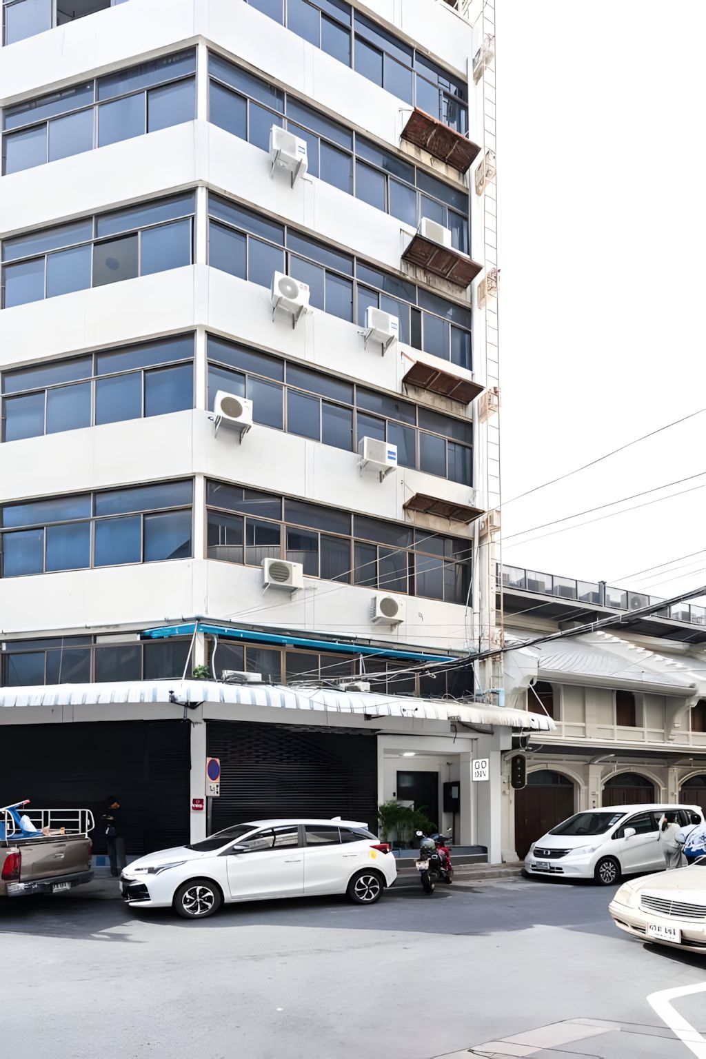 A multi-story building with air conditioning units, a covered parking area, and a street with parked cars and a motorcycle.