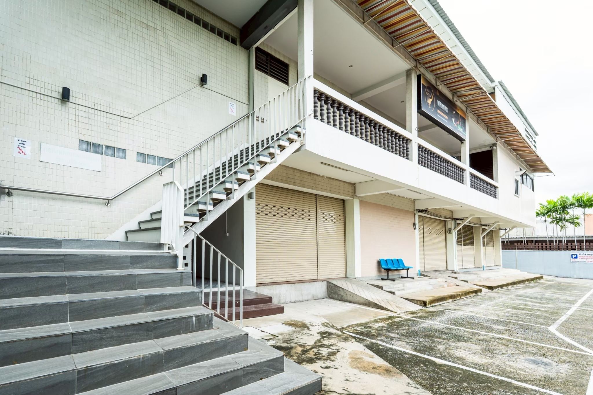 Un edificio de varios pisos con una escalera que lleva a la entrada, un espacio al aire libre techado con bancos y un estacionamiento.