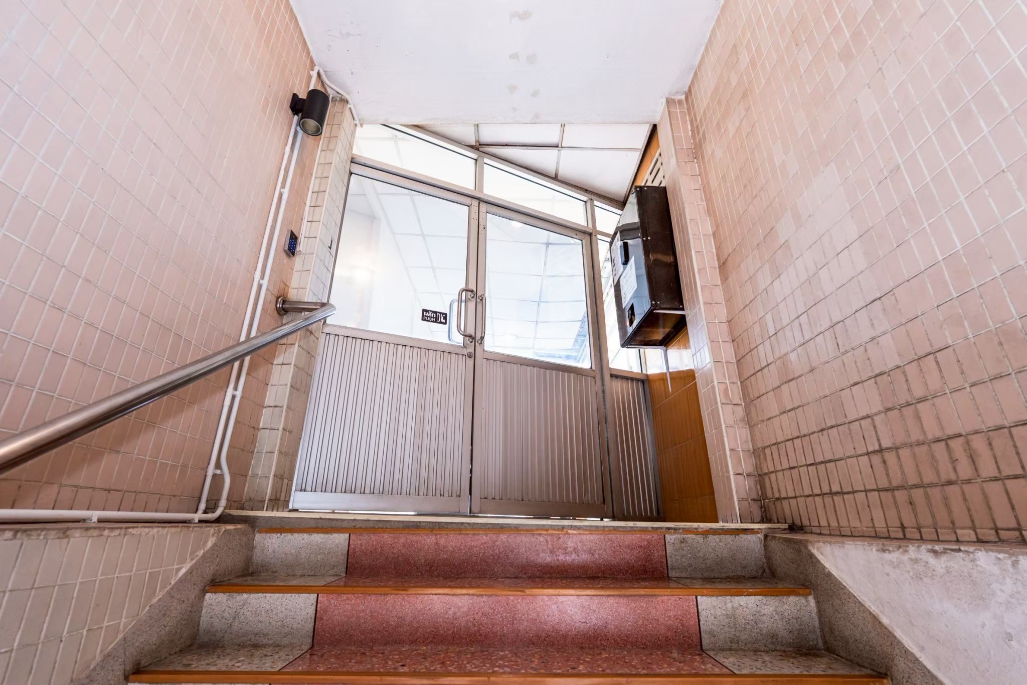 Eine Treppe führt zu einer Tür, an der sich ein Metallgeländer und ein Schild mit der Aufschrift 'Aufzug' befinden.