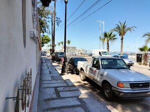 Cena de rua com veículos estacionados, incluindo uma caminhonete branca com 'FESACO' na lateral e uma van branca. Há palmeiras e um céu claro ao fundo.