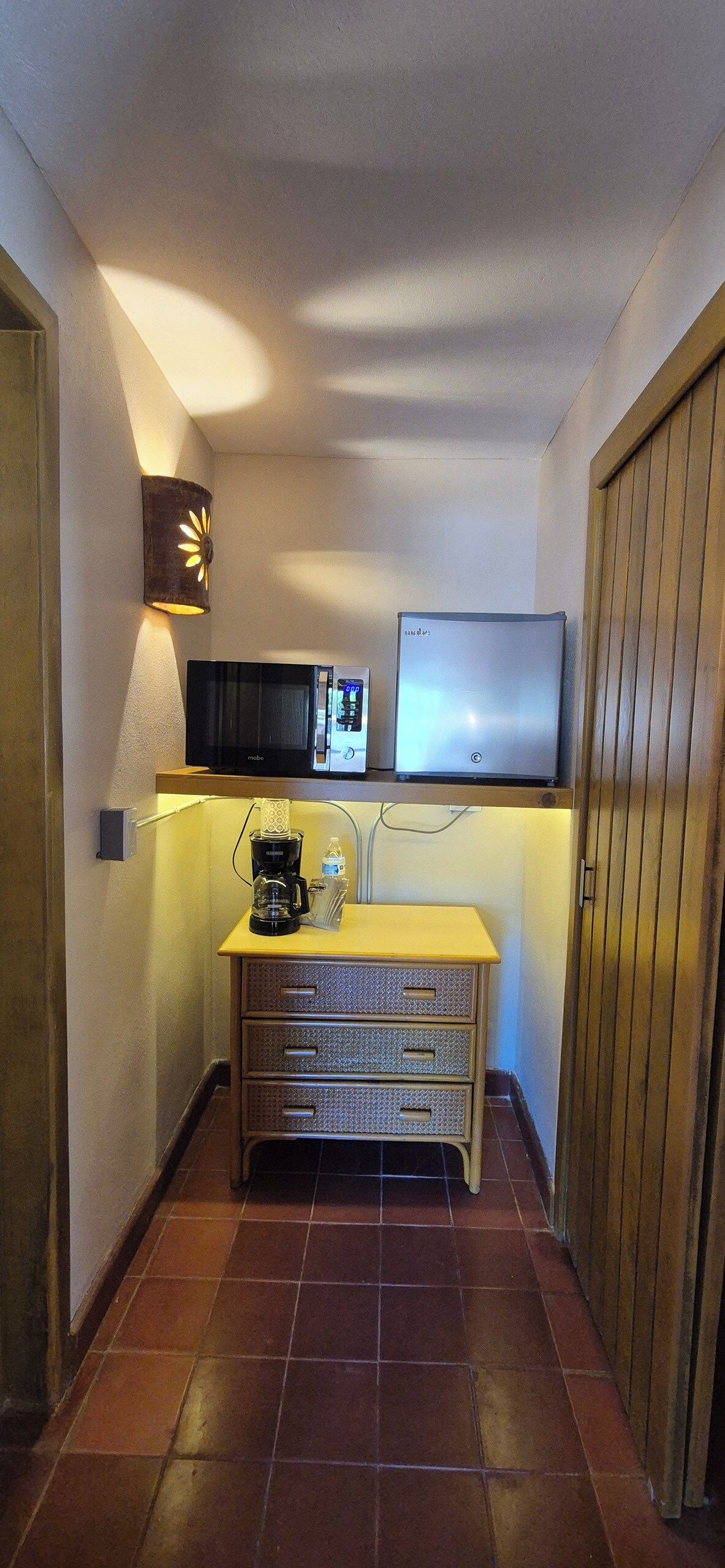 Habitación de hotel compacta con kitchenette, televisión de pantalla plana, lámpara de pared y puerta de madera.