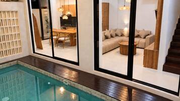 Una piscina interior con terraza de madera, un comedor con mesa y sillas, un sofá y una escalera.