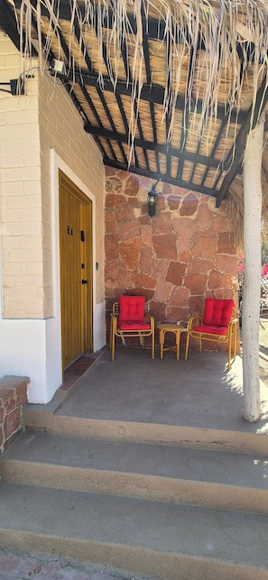 A covered porch with a stone wall, a wooden door, and two wicker chairs with red cushions. - Sandy Shores 8A (La Paz)