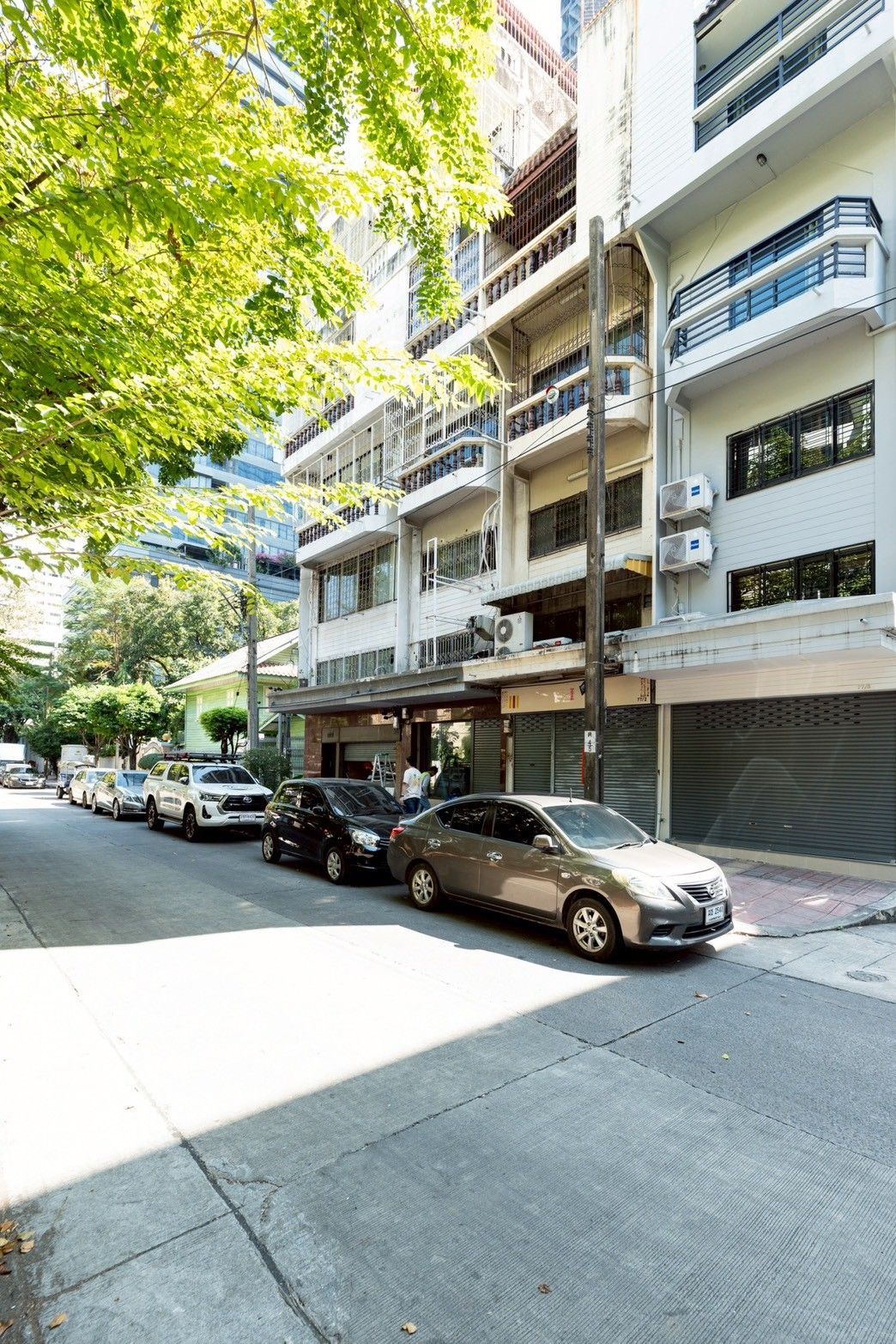 A multi-story residential building with balconies, parked cars, and a sidewalk.