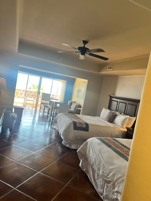 Habitación de hotel con dos camas, ventilador de techo, zona de comedor con mesa y sillas, y vistas al exterior.