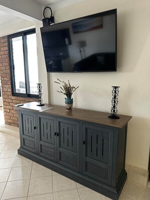 A modern living room with a flat-screen TV mounted on the wall, a dark wooden cabinet with decorative handles, and a potted plant on top. - Cabo Hideaway | Near Beach and Center + Parking. (Cabo San Lucas)