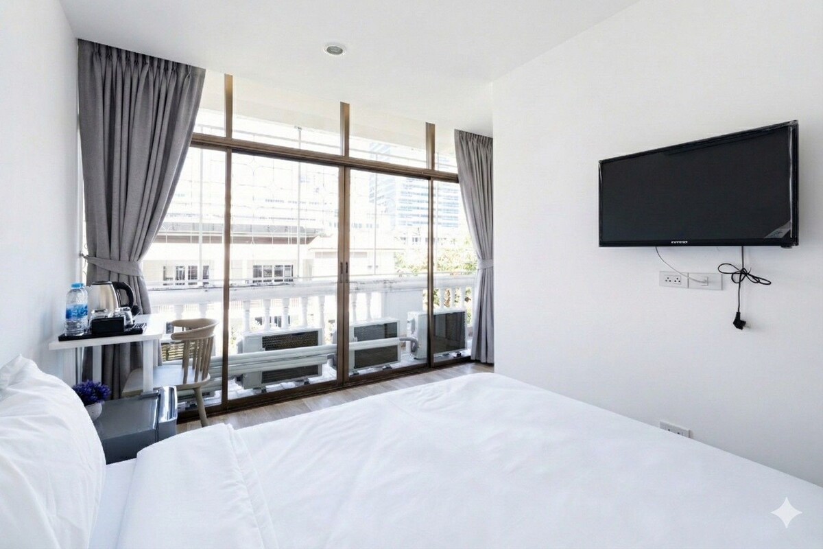 Habitación de hotel con cama, escritorio con tetera, televisión y balcón con vista a edificios.