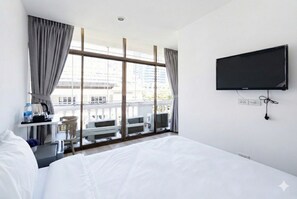 Habitación de hotel con cama, escritorio con tetera, televisión y balcón con vista a edificios.