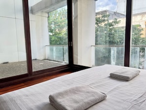 A hotel room with a bed, a window with a view of trees and buildings, and a balcony with a railing. - H room De Silom (Bangkok)