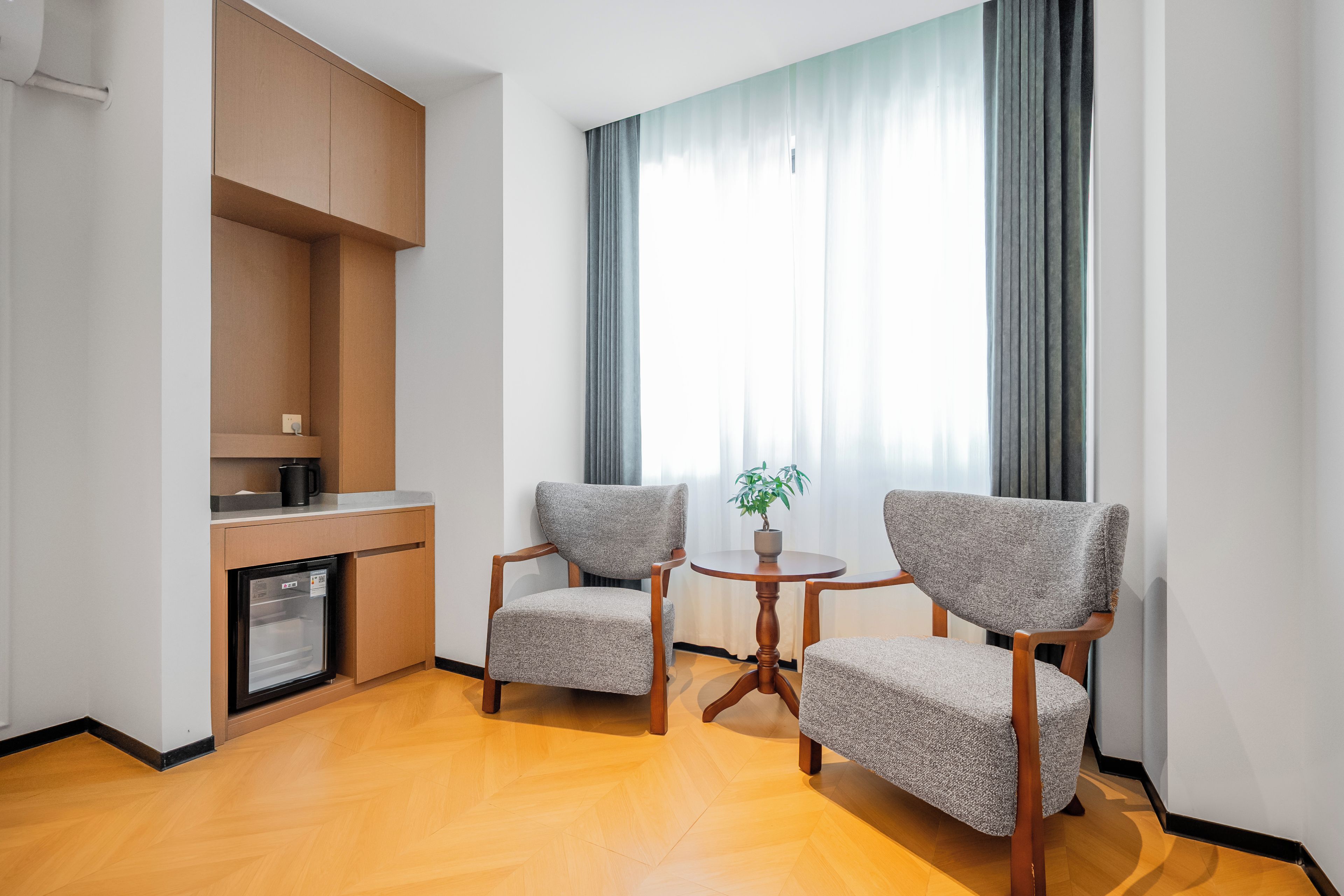 A modern hotel room with a kitchenette, two armchairs, a small round table, and a large window with curtains.