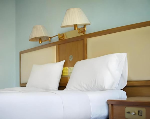 Habitación de hotel con cabecera de madera, dos lámparas de noche, un reloj y una mesita de noche con cajón.