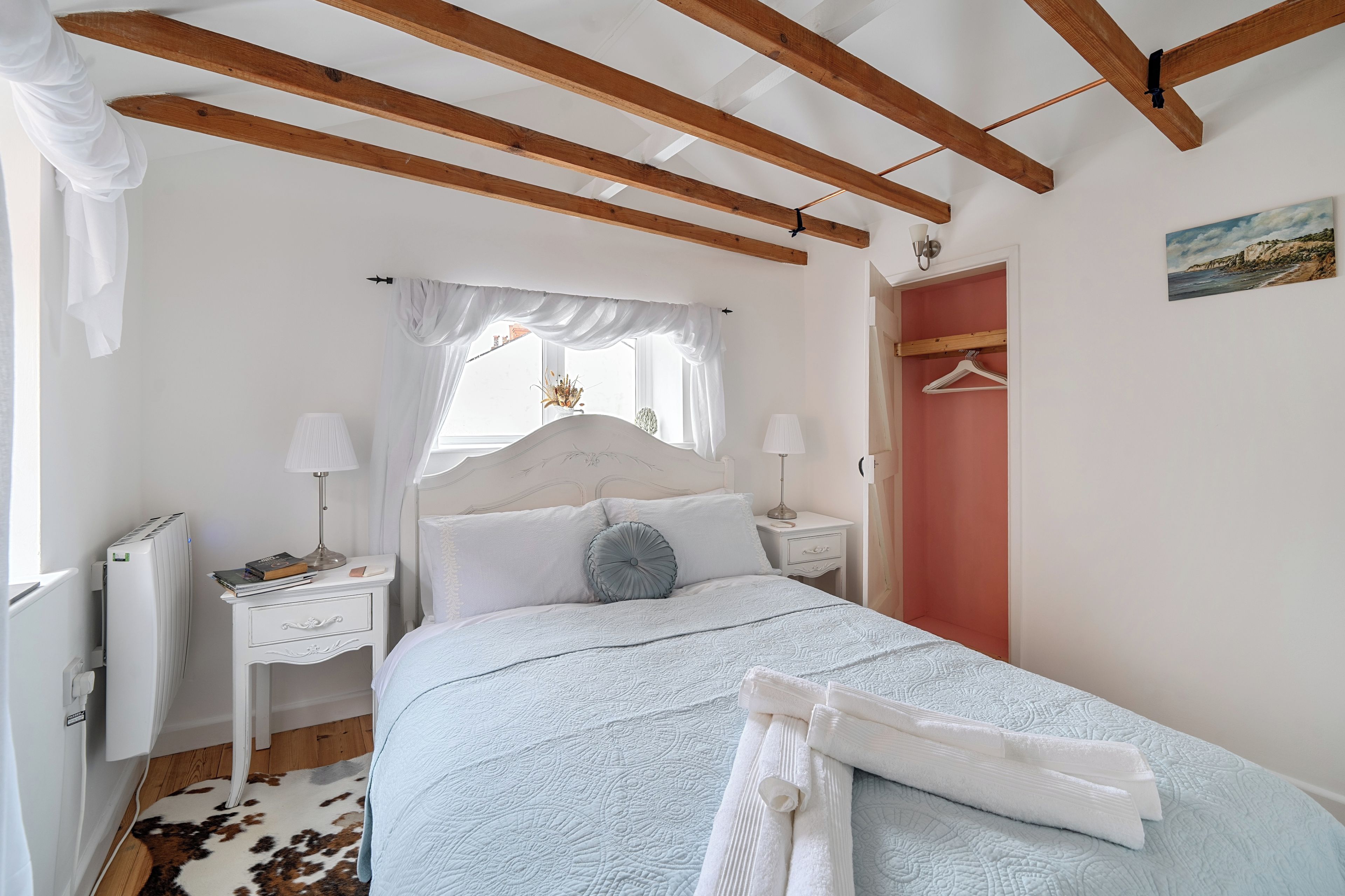 A bedroom with a bed, bedside tables, a window with curtains, a radiator, a coat rack, and a painting on the wall.