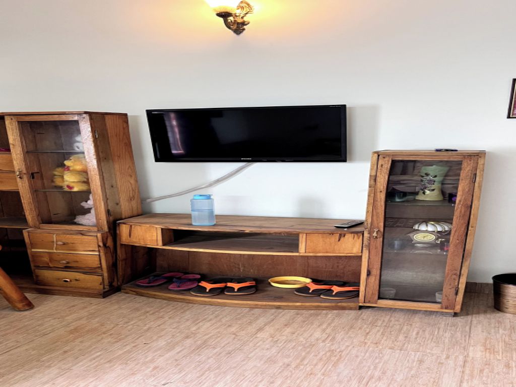 A room with a flat-screen TV mounted on the wall, wooden furniture including a cabinet and a shelf, and a pair of flip-flops on the floor.