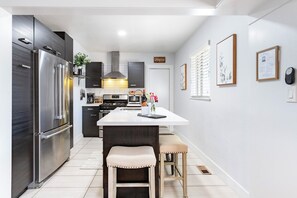 A modern kitchen with a stainless steel refrigerator, dark cabinetry, a central island with seating, and a window with blinds. - West Highlands- walk to Tennyson and Highlands Square (Denver)