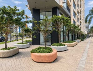 A modern building with a paved walkway, surrounded by landscaped planters with small trees. - Elegante 2BR con piscina y vistas (Miraflores)