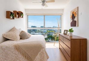 A bedroom with a bed, a dresser, a ceiling fan, a window with a city view, and wall-mounted shelves with plants. - Elegante 2BR con piscina y vistas (Miraflores)