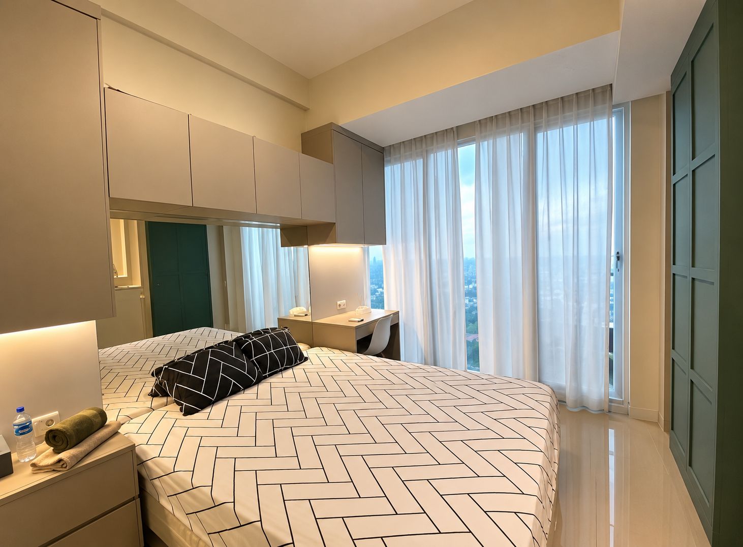 Une chambre d’hôtel moderne avec un grand lit, une table de chevet avec une lampe, une vue sur la ville grâce à de grandes fenêtres et une porte verte.