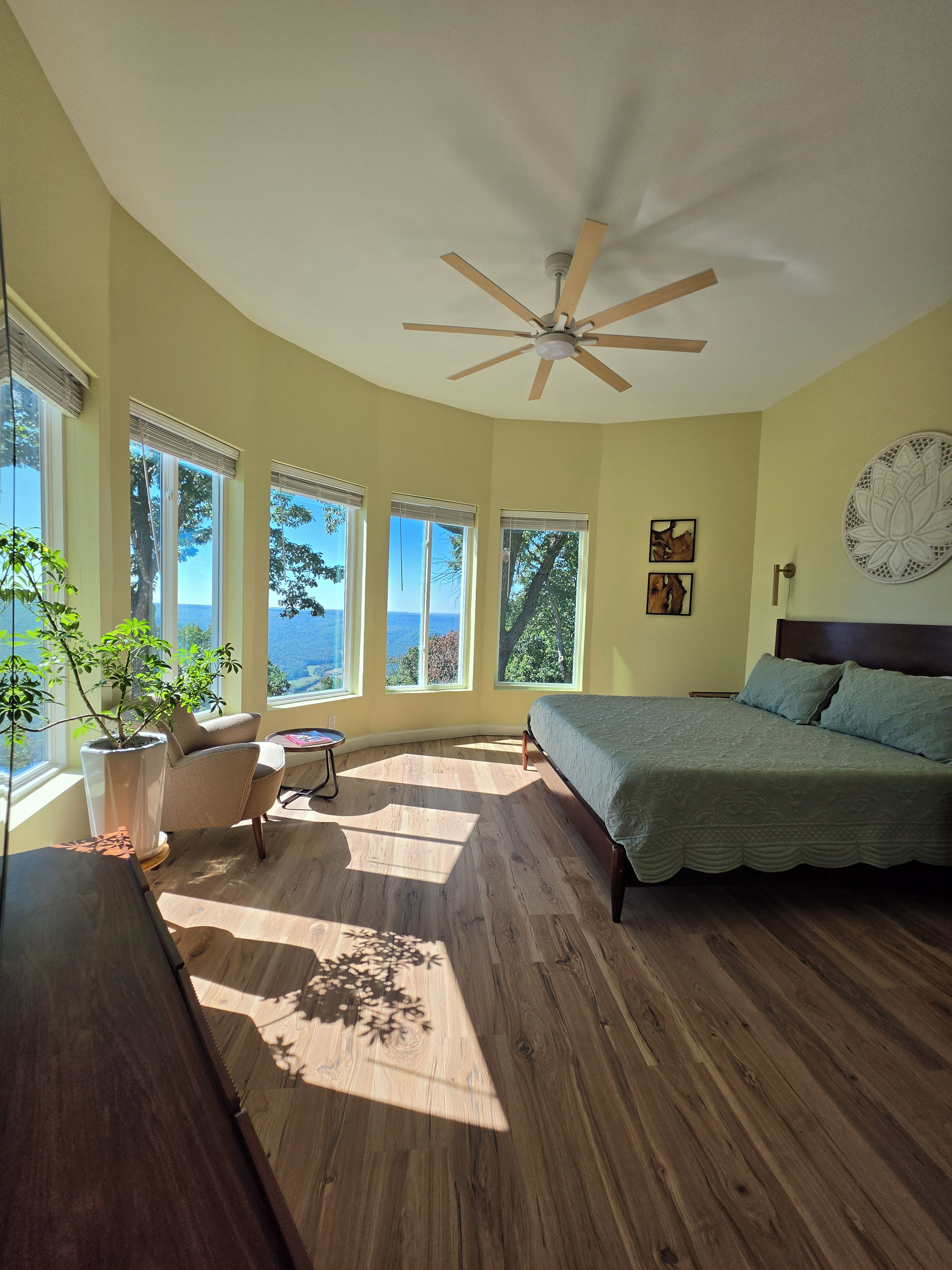 Une chambre à coucher avec un lit, un ventilateur de plafond, une plante en pot et de grandes fenêtres donnant sur un beau paysage.