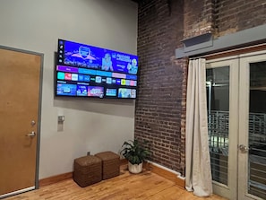 Rebedor d'hotel amb un televisor de pantalla plana gegant fixat a la paret que mostra diversos canals i un partit esportiu. Hi ha dos otomans marrons i una planta en maceta al terra.